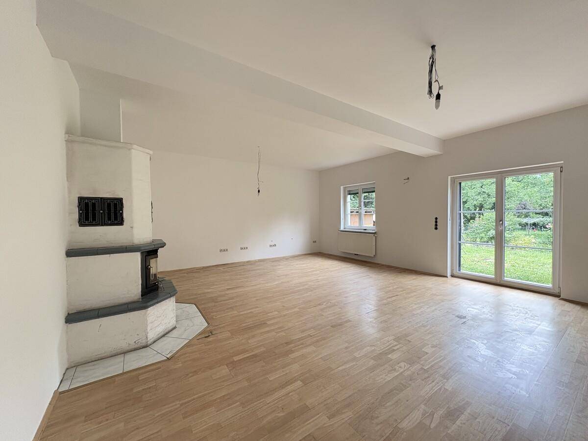 Geräumiges Wohnzimmer mit hellem Parkettboden, Kachelofen und Zugang zum Garten.