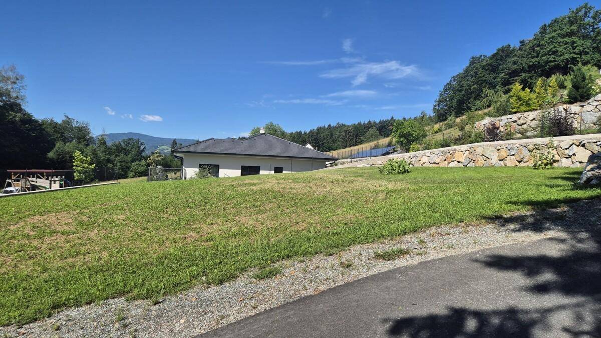 Weitläufige Rasenfläche mit einem modernen Haus im Hintergrund und einer Natursteinmauer am Hang.