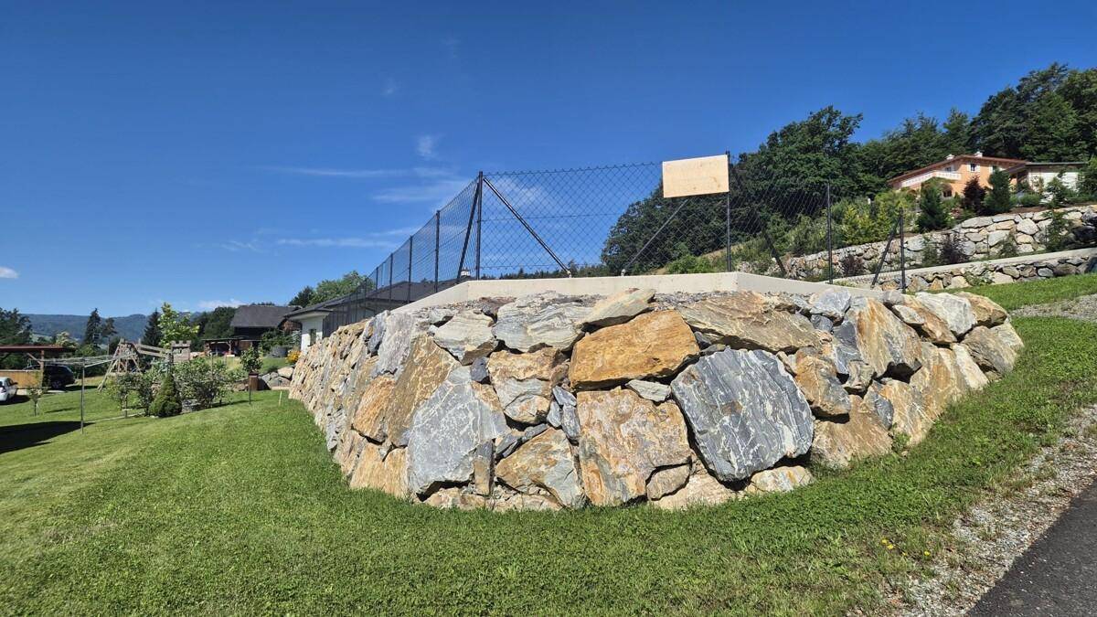 Massive Natursteinmauer mit Zaun als Grundstücksbegrenzung, umgeben von Grünflächen und Häusern.
