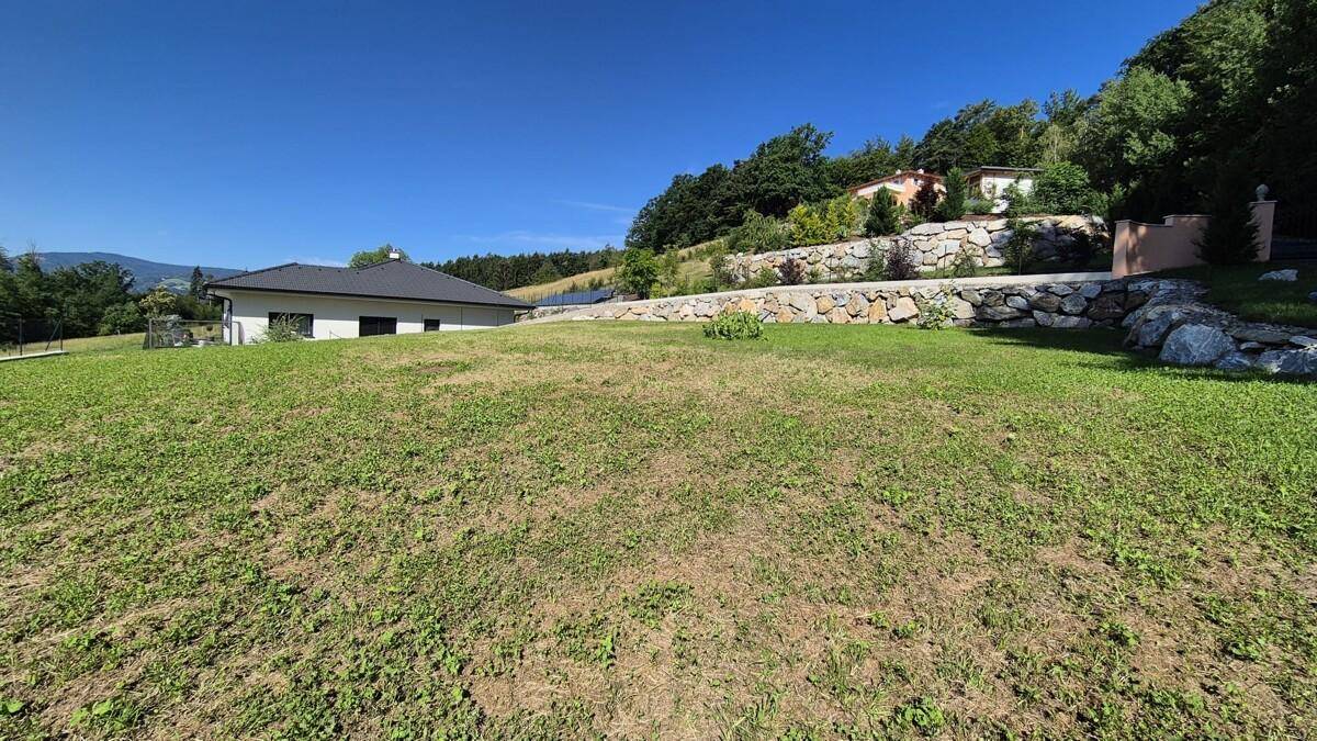 Große, sonnige Rasenfläche mit einem modernen Haus und einer markanten Natursteinmauer im Hintergrund.