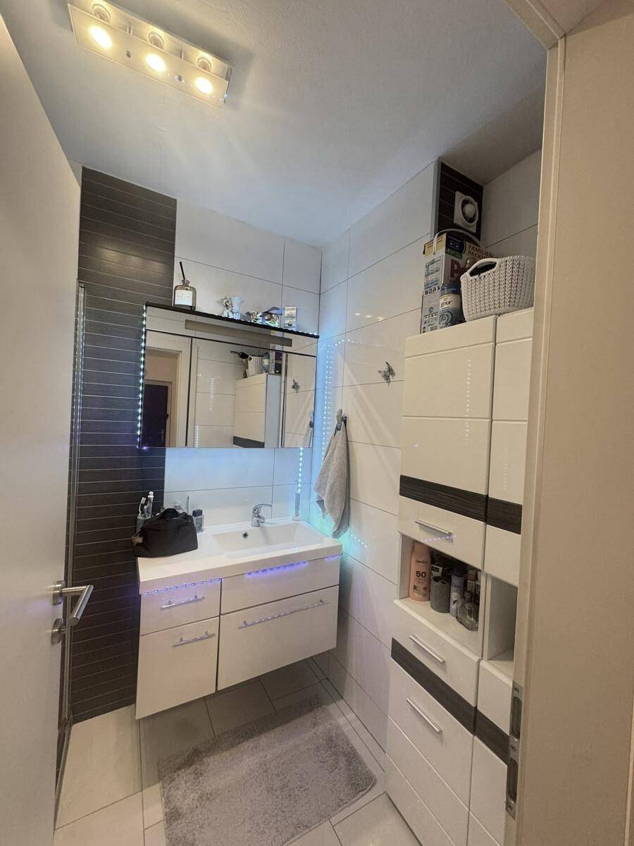 Modernes Badezimmer mit stilvollem Waschtisch, beleuchtetem Spiegelschrank und praktischen Hochschränken. Weiße Fliesen und dunkle Akzentwand schaffen eine zeitgemäße Atmosphäre.