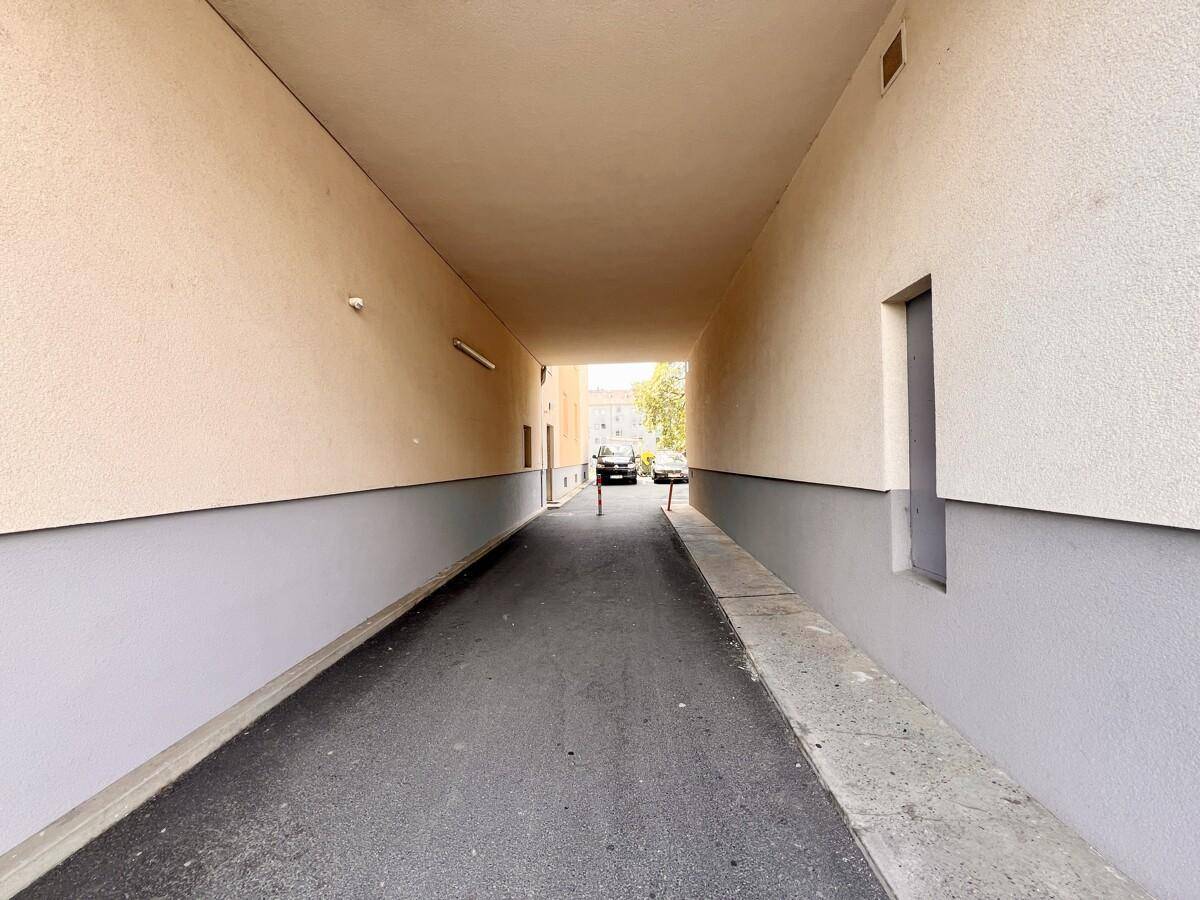 Überdachter Durchgang mit beigen Wänden und grauen Sockeln, der zu einem Parkplatz führt.