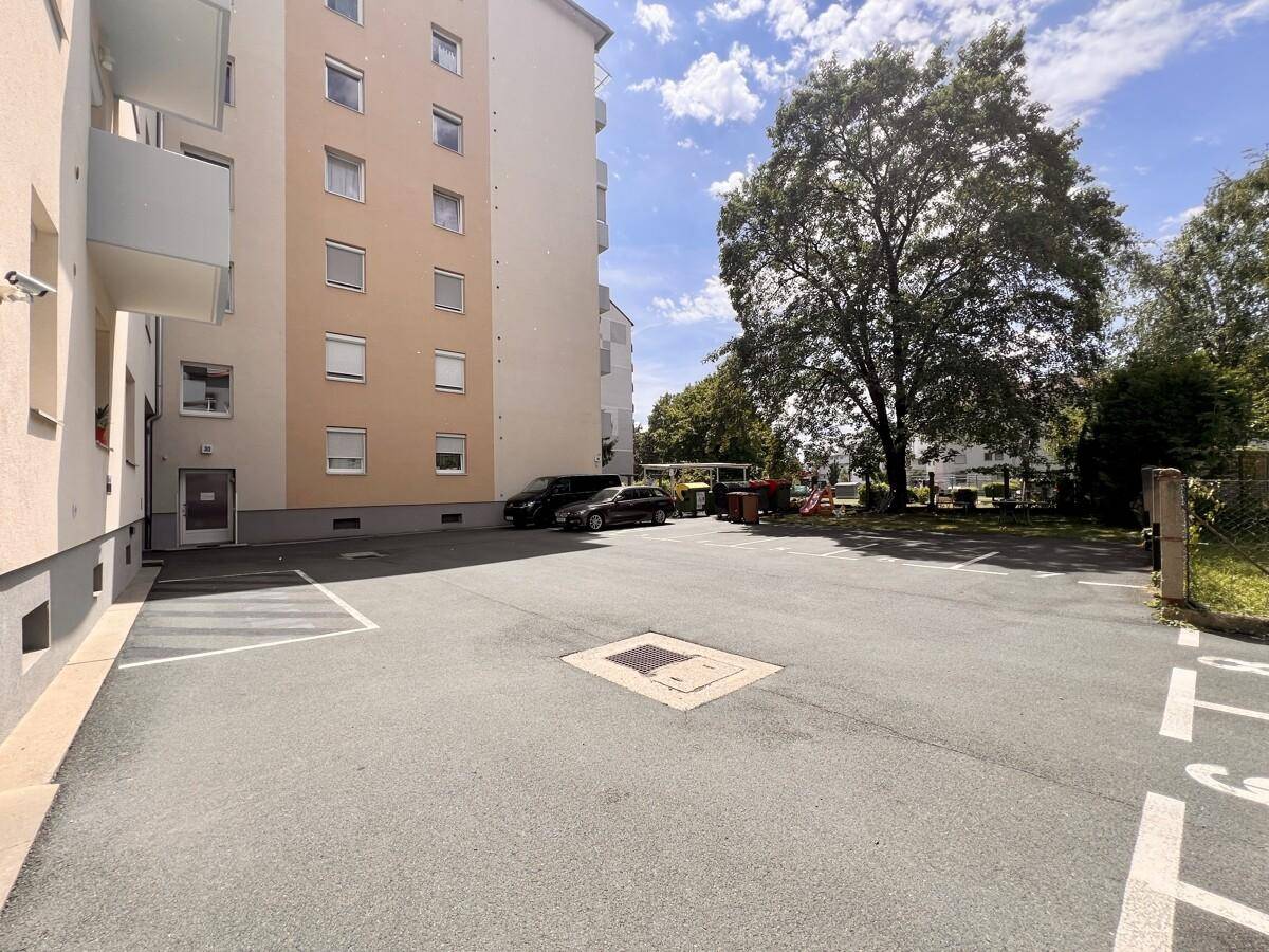 Außenparkplatz vor einem Mehrfamilienhaus mit Bäumen und Grünflächen im Hintergrund.