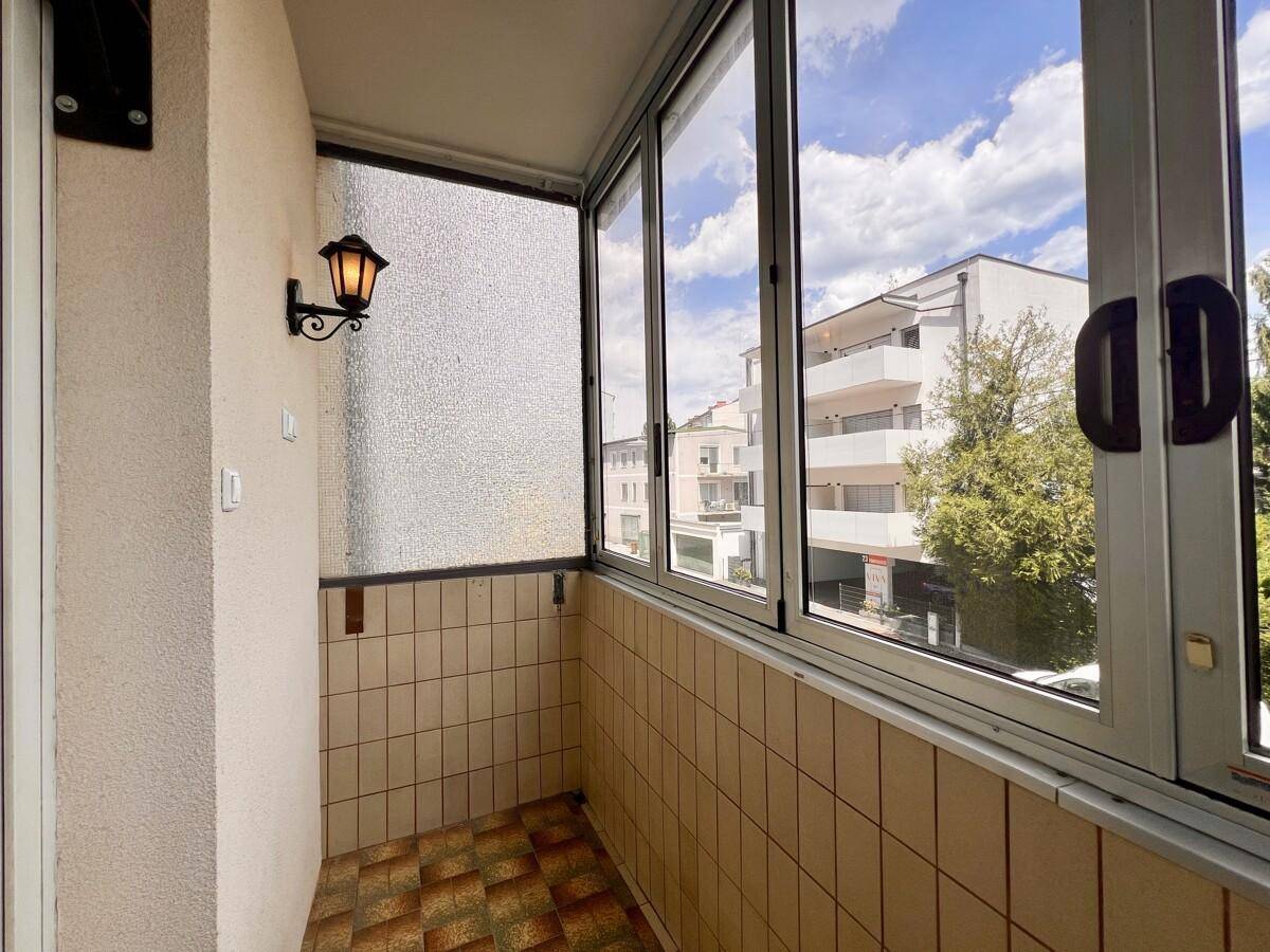 Verglaste Loggia mit Fliesenboden und Blick auf die umliegenden Gebäude und Bäume.