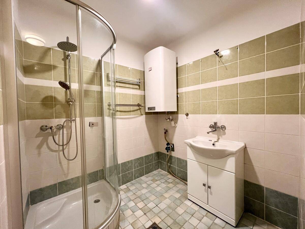 Badezimmer mit Duschkabine, Waschbecken und Boiler, gefliest in Grün- und Beigetönen.