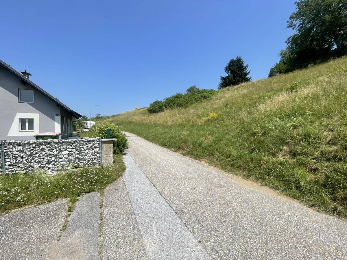 Straße neben einem Haus mit Gabionenwand und einem grünen, sonnigen Hang.