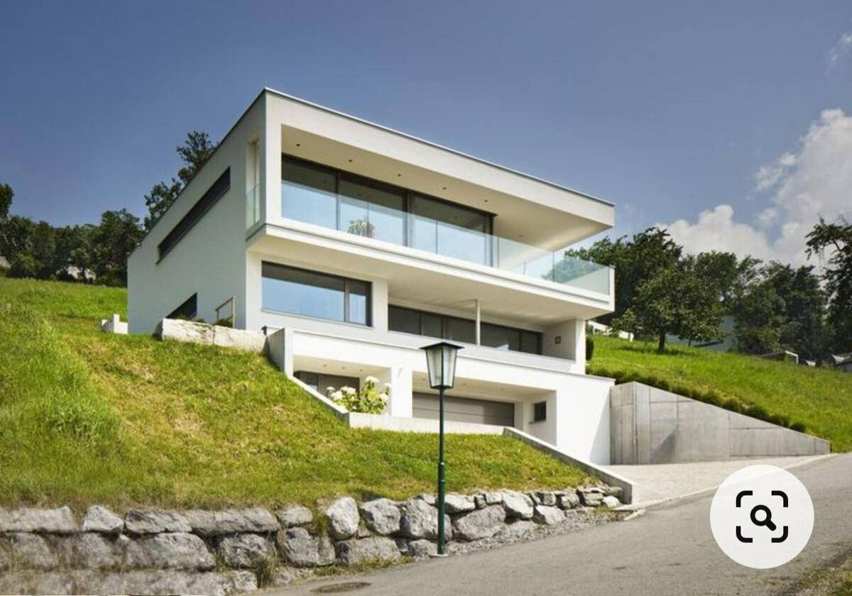 Modernes, weißes Haus mit großen Glasfronten, elegant in einen grünen Hang integriert.