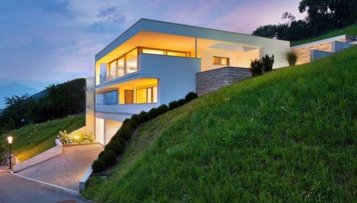 Modernes Haus am Hang bei Dämmerung, mit warm beleuchteten Innenräumen und großen Fenstern.