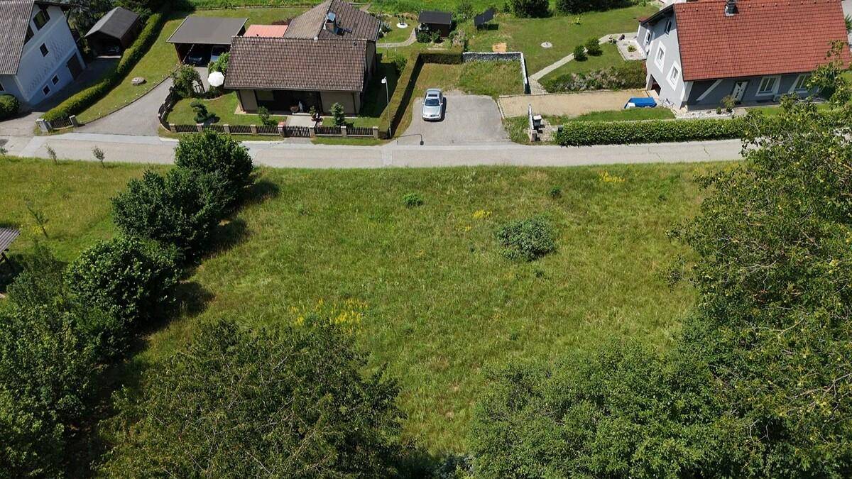 Luftaufnahme eines unbebauten, grünen Grundstücks mit angrenzender Straße und Wohnhäusern.