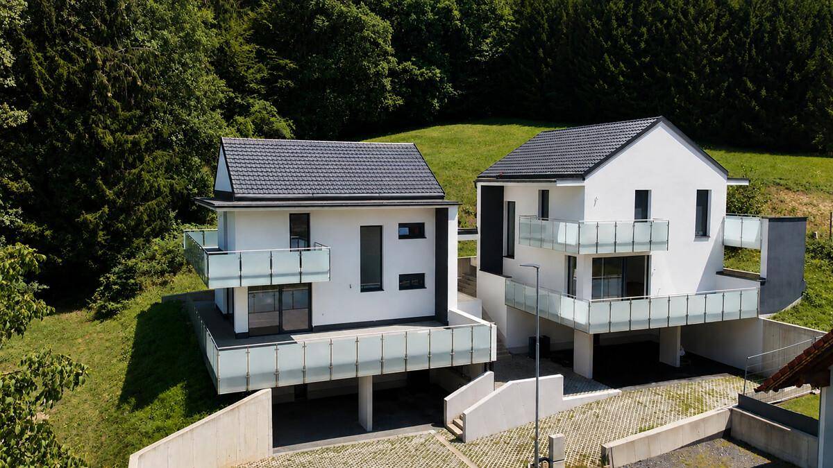 Zwei moderne Häuser mit dunklen Dächern und Glasbalkonen in ländlicher Lage.
