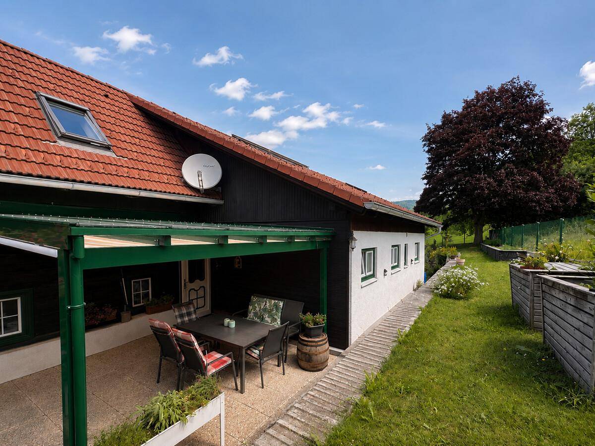 Hausfassade mit überdachter Terrasse, Garten und Blick auf die umliegende Landschaft.