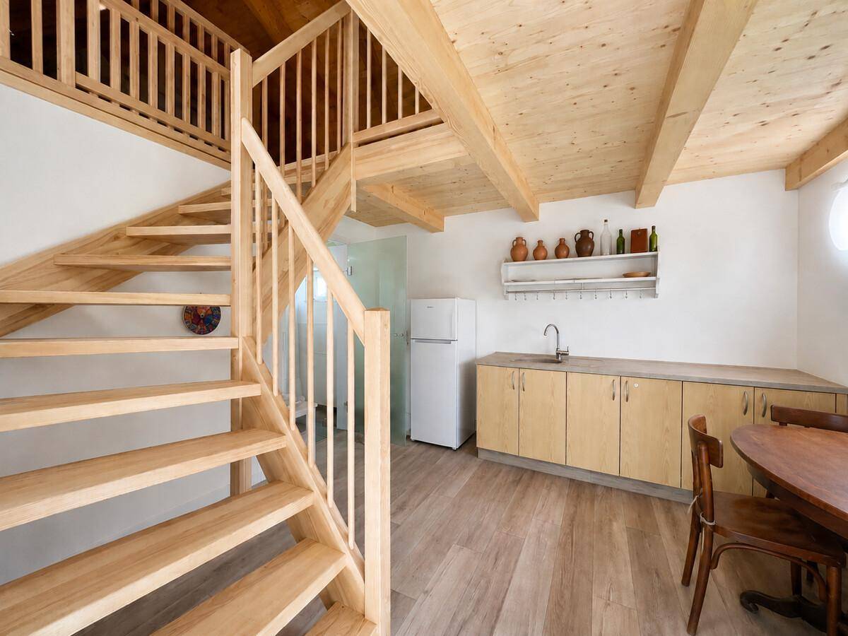 Heller Wohnbereich mit offener Holztreppe, Holzbalkendecke und kleiner Küchenzeile.