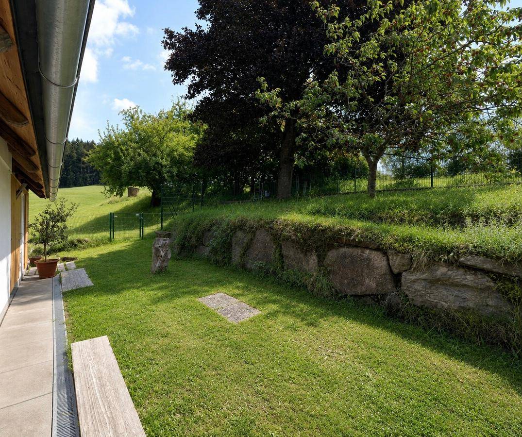 Sonniger Gartenbereich mit Rasenfläche, alter Steinmauer und schattenspendenden Bäumen.