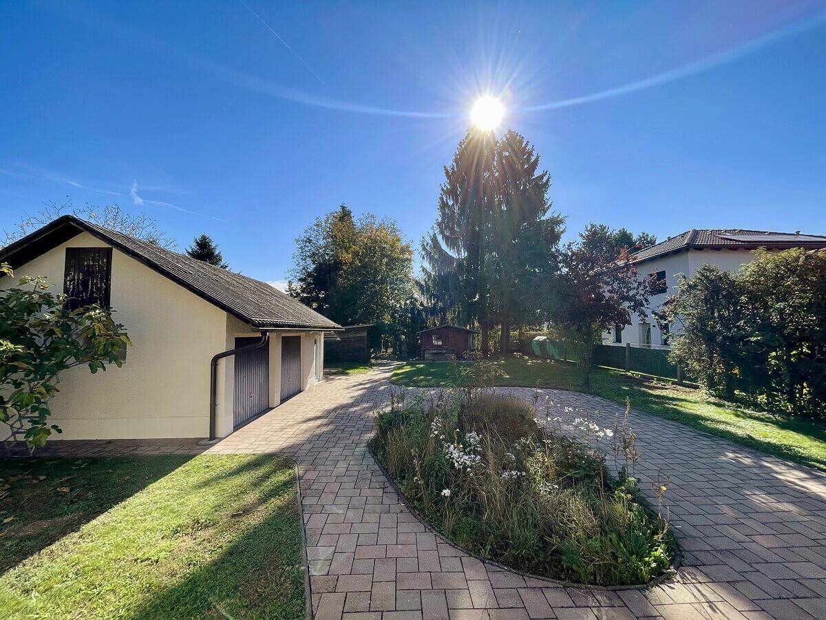 Sonnige Außenansicht mit Garagengebäude, gepflasterter Auffahrt und grünem Gartenbereich.