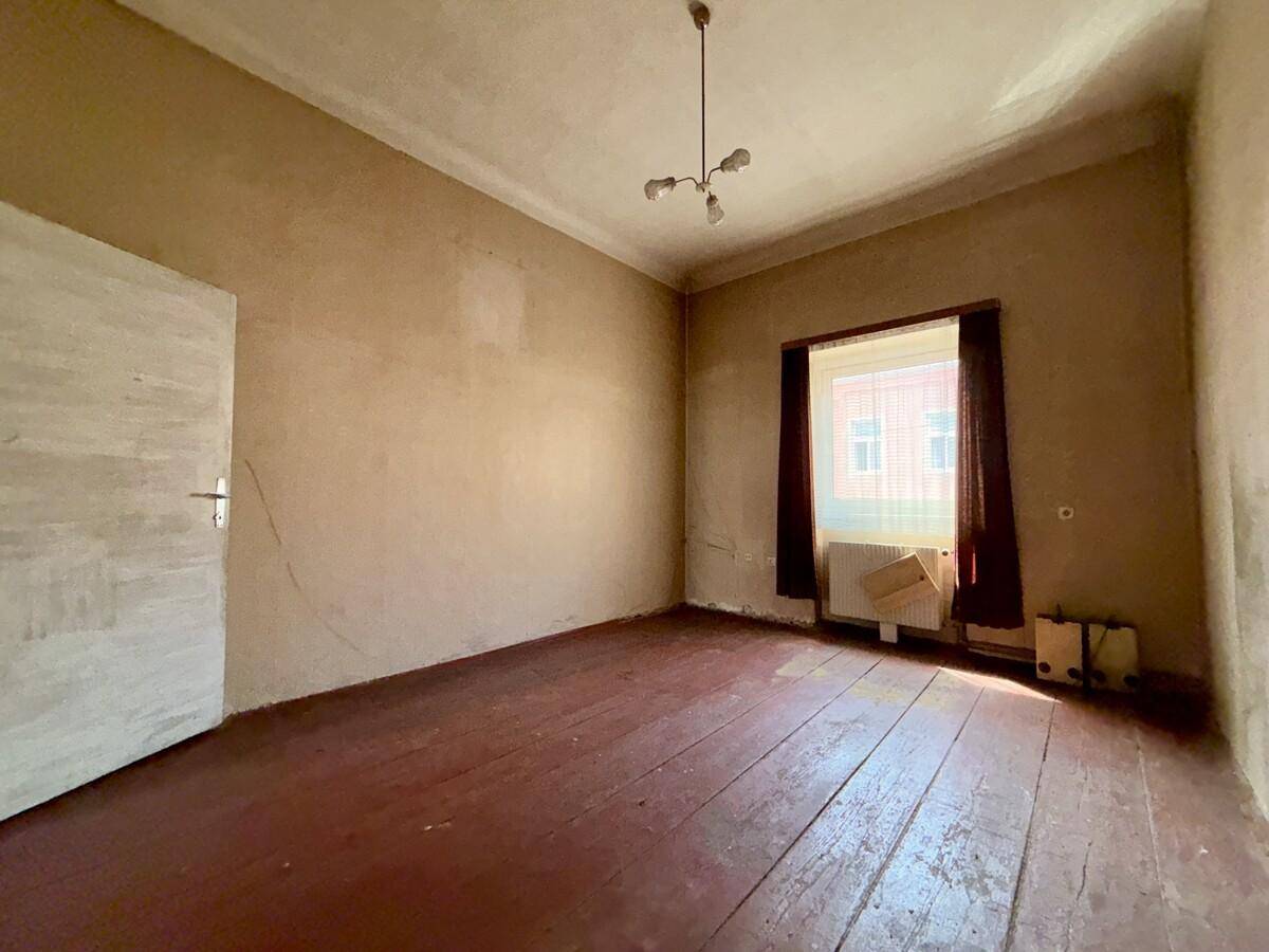 Leeres Zimmer mit abgenutztem Holzboden, einfacher Deckenleuchte und einem Fenster zur Straße.