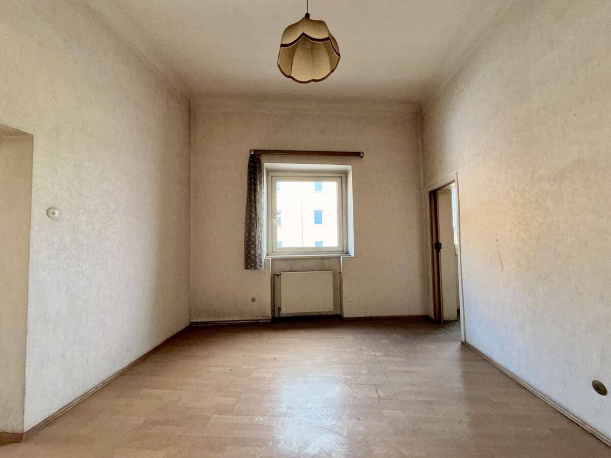 Leeres Zimmer mit alter Tapete, einem Fenster und einer einfachen Deckenlampe.