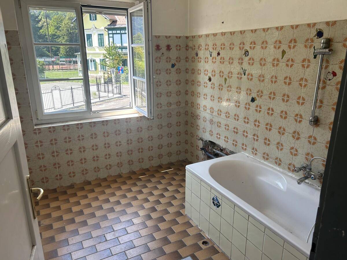 Badezimmer mit älteren Fliesen, Badewanne und Fenster, das viel Tageslicht hereinlässt.