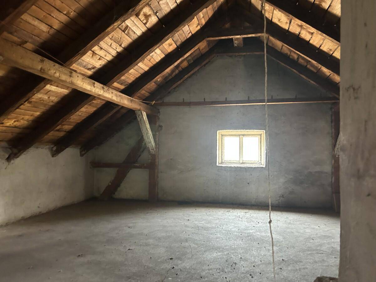Unausgebauter Dachboden mit sichtbaren Holzbalken, Betonboden und einem kleinen Fenster.