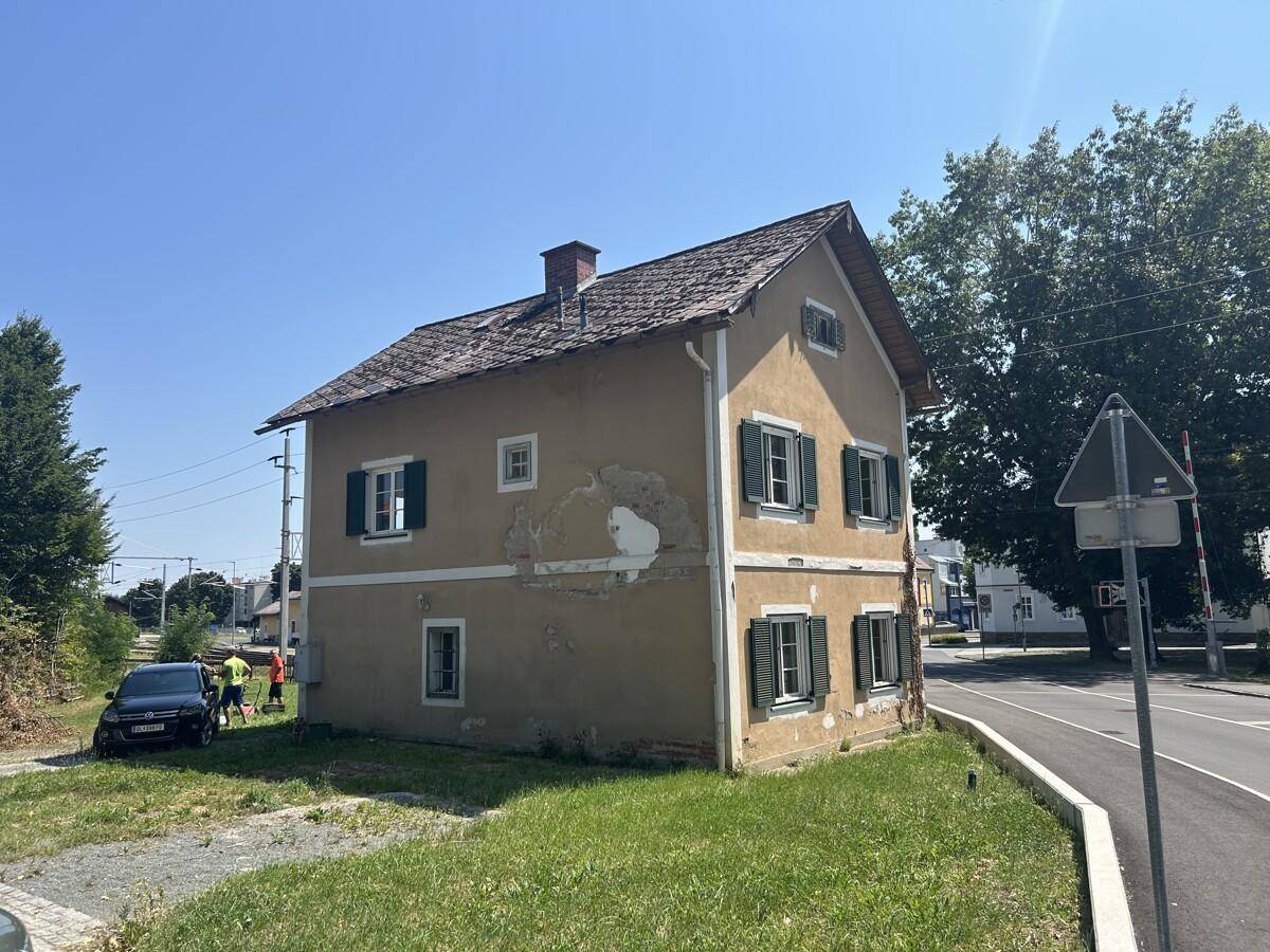 Ansicht des alten Hauses mit abblätterndem Putz, grünen Fensterläden und angrenzender Straße.