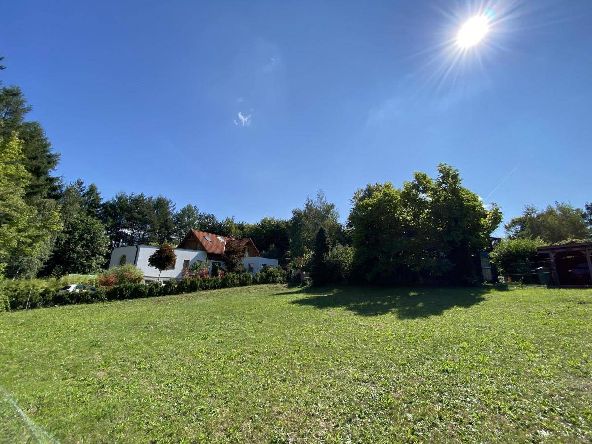 Sonnige Rasenfläche mit Bäumen und einem Wohnhaus im Hintergrund, umgeben von Natur.