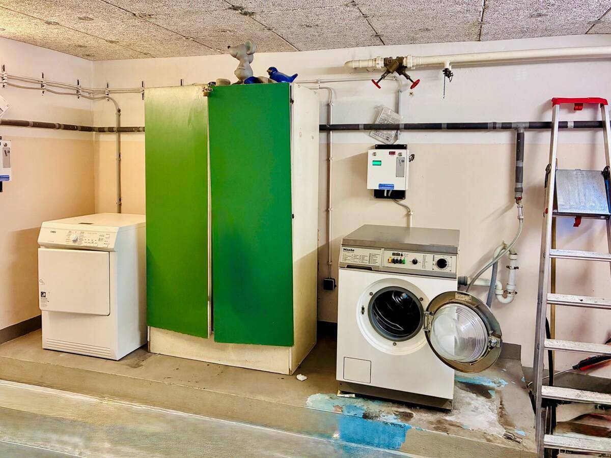 Funktionaler Waschraum im Keller mit Waschmaschine, Trockner und sichtbaren Rohrleitungen.