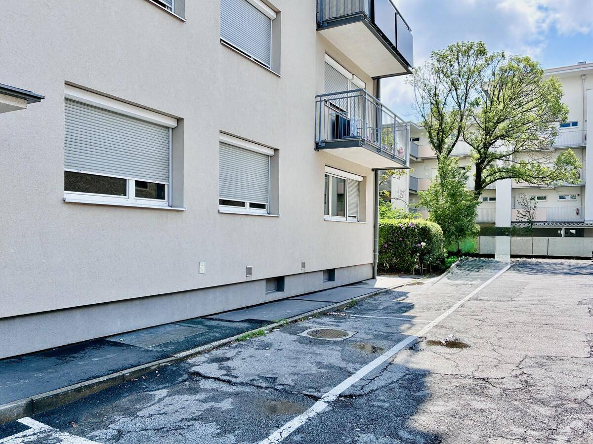 Außenansicht eines Mehrfamilienhauses mit Balkonen, Rollläden und Parkplätzen im Freien.