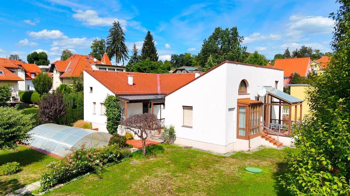 Ansicht des Hauses von der Gartenseite mit großem Rasen, überdachtem Pool und sonnigem Himmel.