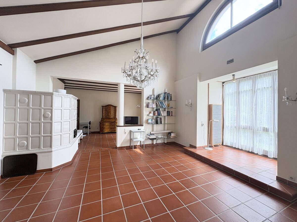 Geräumiges Wohnzimmer mit hohen Decken, sichtbaren Holzbalken, Terrakottafliesen und einem Kachelofen.