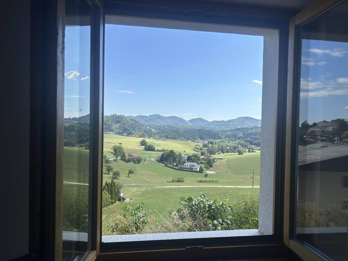 Fensterblick auf eine weite, grüne Hügellandschaft mit vereinzelten Häusern.