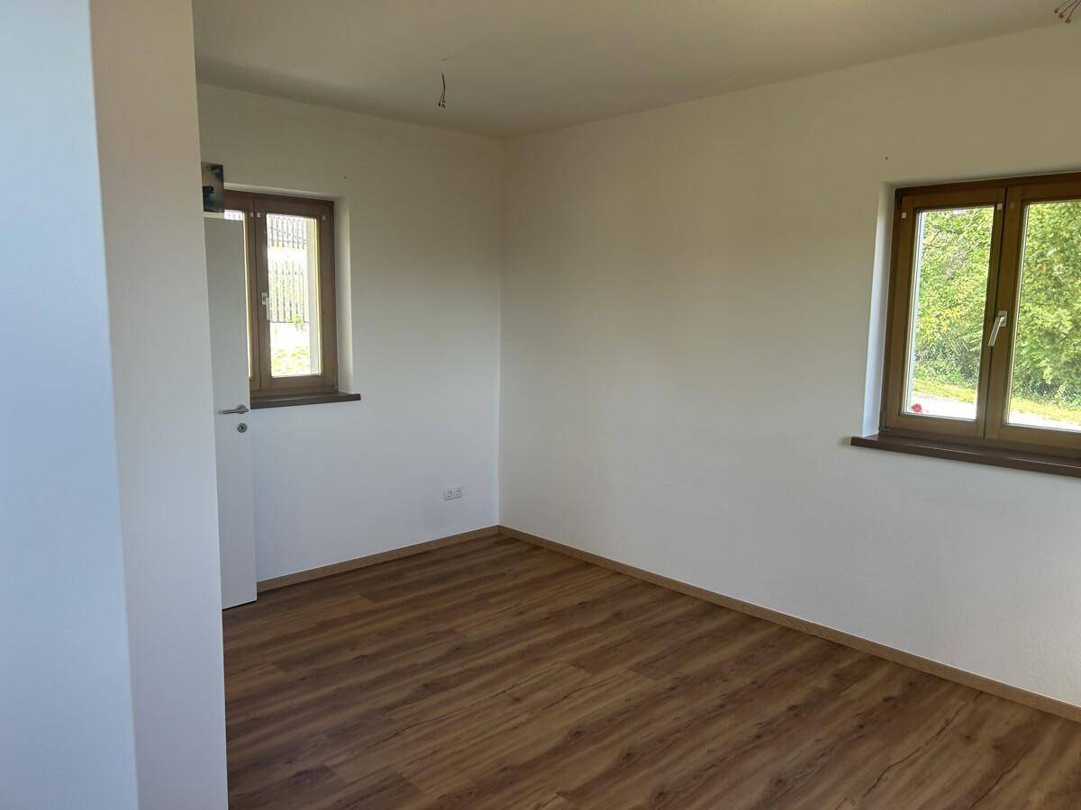 Helles Zimmer mit Laminatboden und zwei Fenstern, die den Blick ins Grüne freigeben.