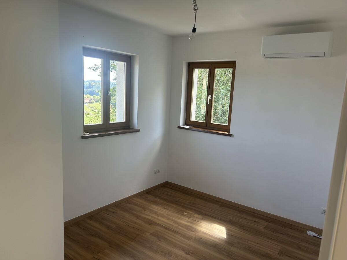 Helles Zimmer mit Laminatboden, zwei Fenstern und einer Klimaanlage an der Wand.