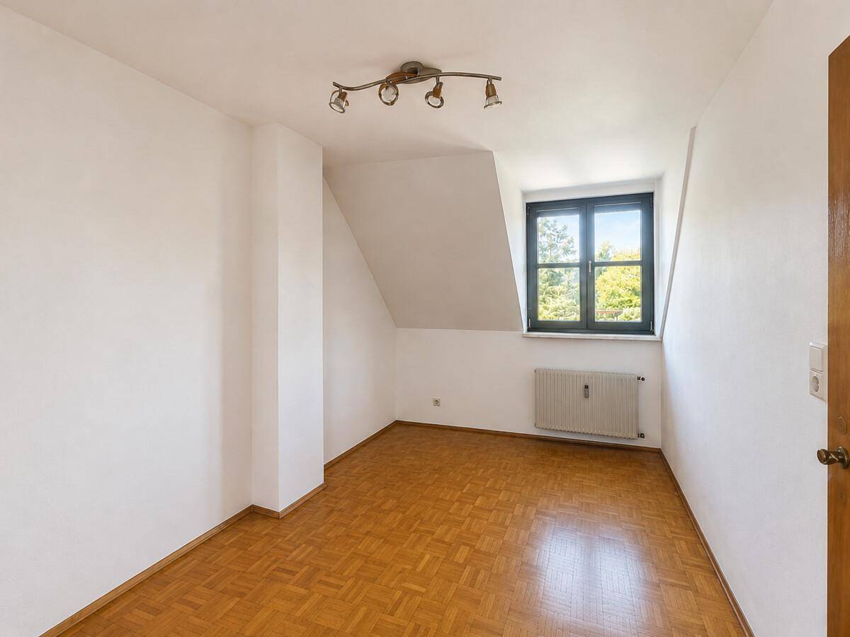 Freundliches Zimmer mit Holzparkett, schräger Decke und einem Fenster mit Ausblick ins Grüne.