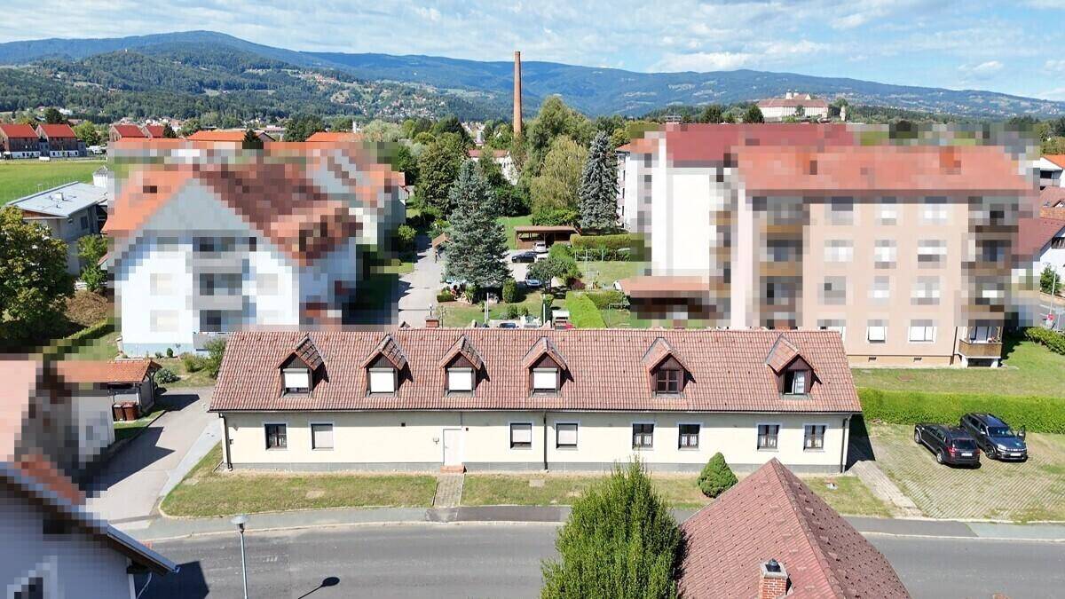 Luftaufnahme des Gebäudes mit Gaubenfenstern, umgeben von Wohnbebauung und einer bergigen Landschaft im Hintergrund.