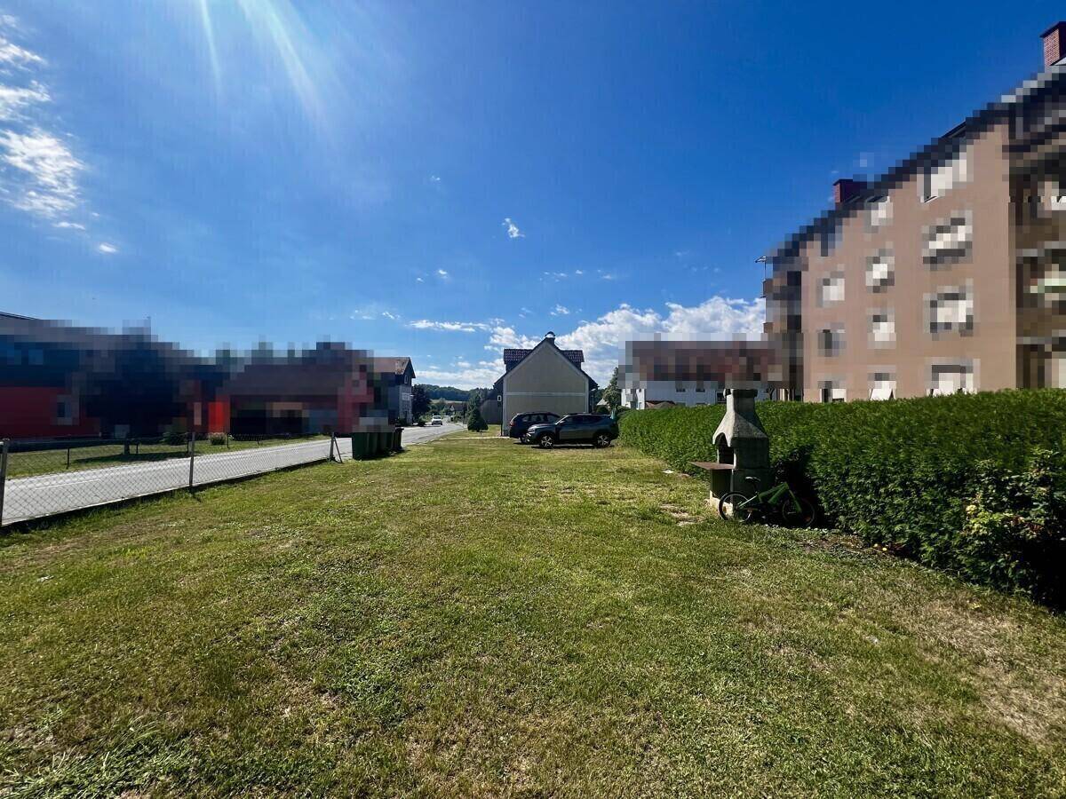 Große Rasenfläche mit Hecke und Grillplatz, umgeben von weiteren Gebäuden unter blauem Himmel.
