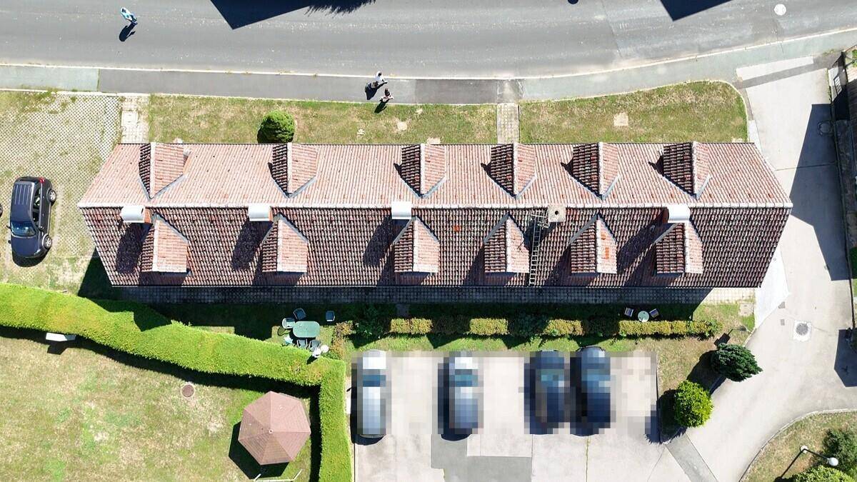 Draufsicht auf das Gebäude mit Ziegeldach und Gauben, umgeben von Parkplätzen und einem möblierten Gartenbereich.