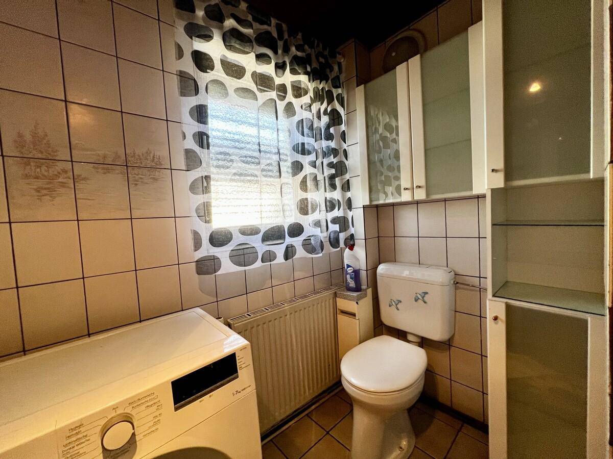 Badezimmer mit Toilette, Waschmaschine und Fenster, ausgestattet mit hellen Fliesen.
