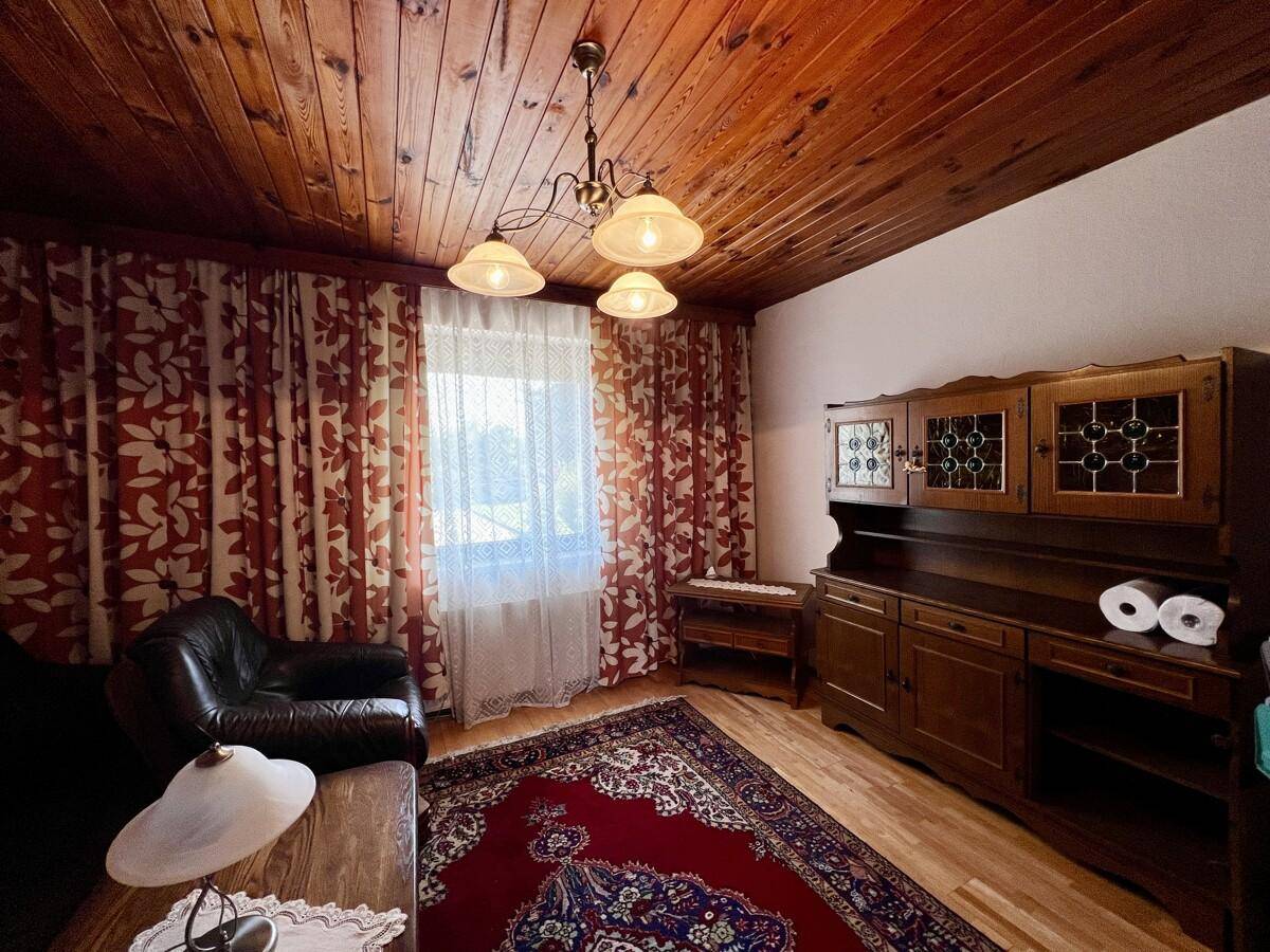 Wohnzimmer mit großem Holzschrank, Fenster und traditionellem Teppich auf Parkettboden.