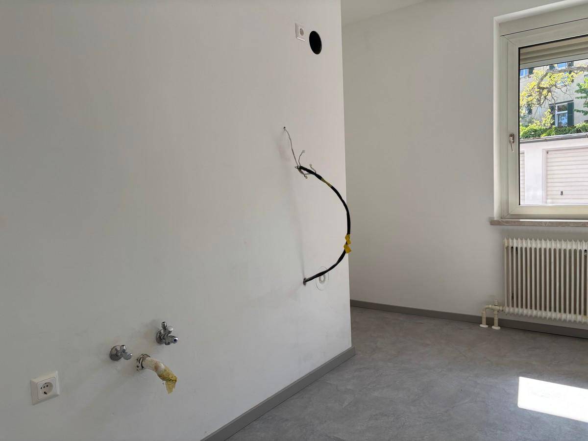 Leerer Küchenraum mit Anschlüssen für Spüle und Geräte, hellen Wänden und grauem Boden.