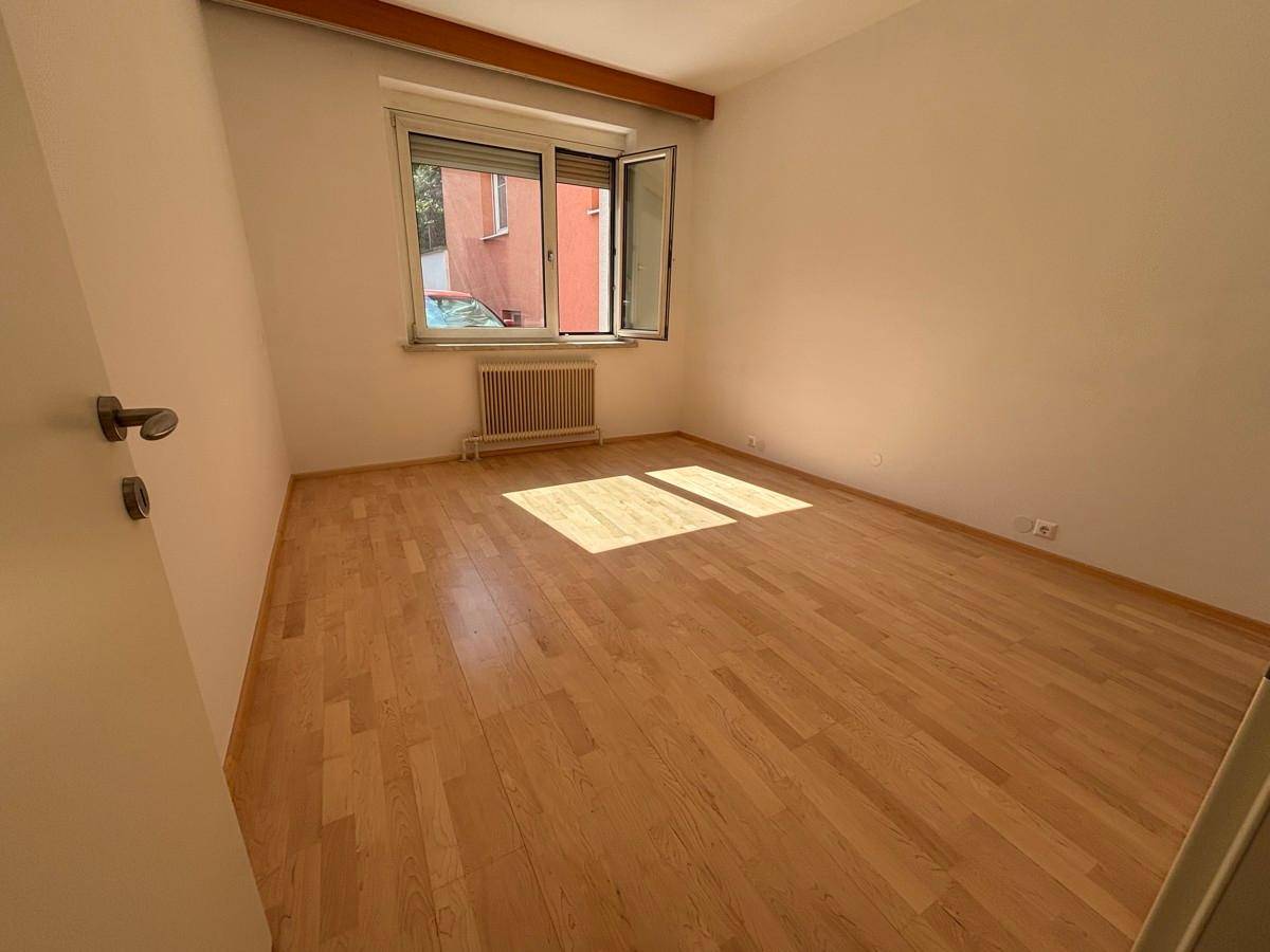 Helles, leeres Zimmer mit Holzparkettboden, weißen Wänden und einem Fenster mit Heizkörper.
