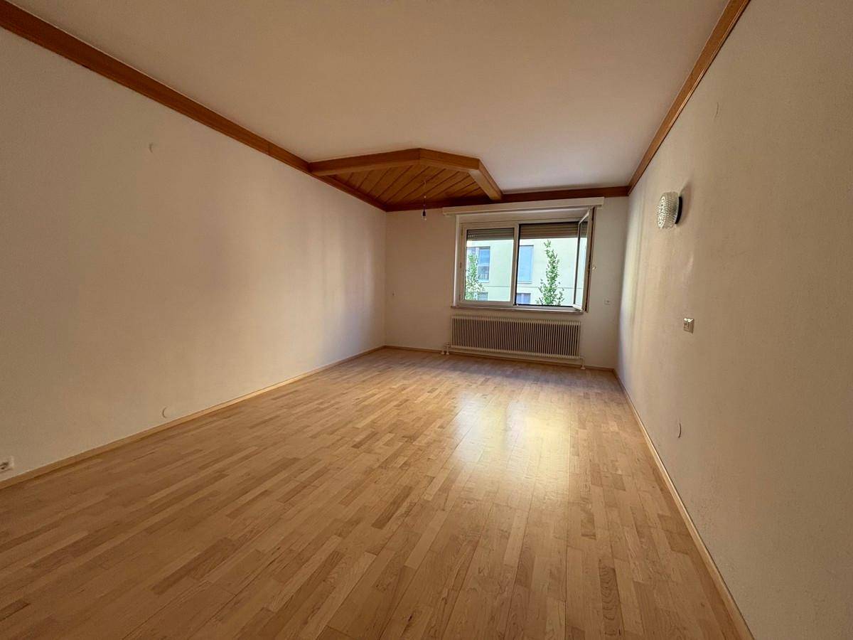 Geräumiges, leeres Wohnzimmer mit Holzparkettboden, weißen Wänden und dekorativen Holzdeckenbalken.