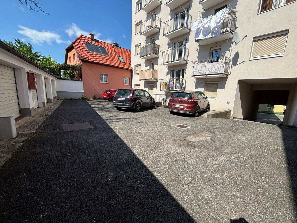 Hinterhofbereich eines Mehrfamilienhauses mit Parkplätzen und Garagen unter blauem Himmel.