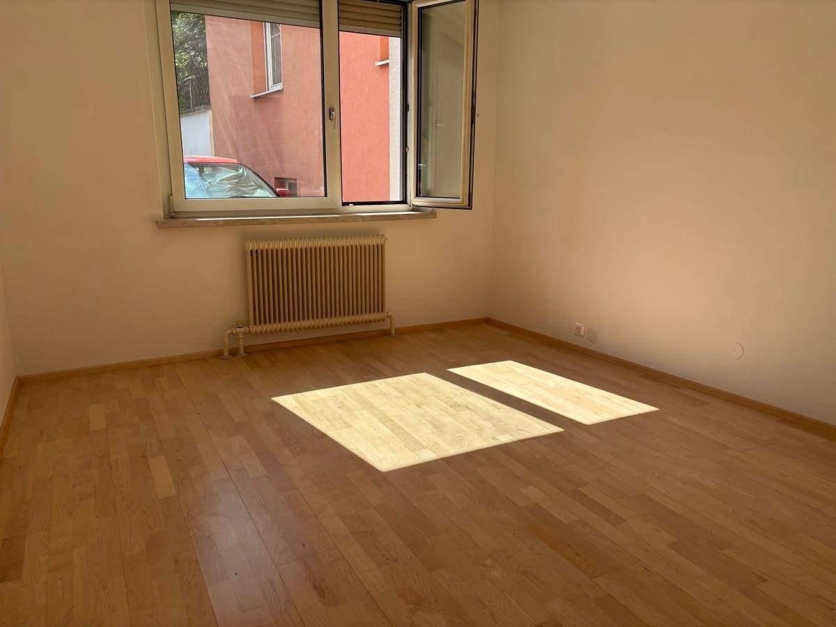 Helles, leeres Zimmer mit Holzparkettboden, weißen Wänden und einem Fenster mit Heizkörper.