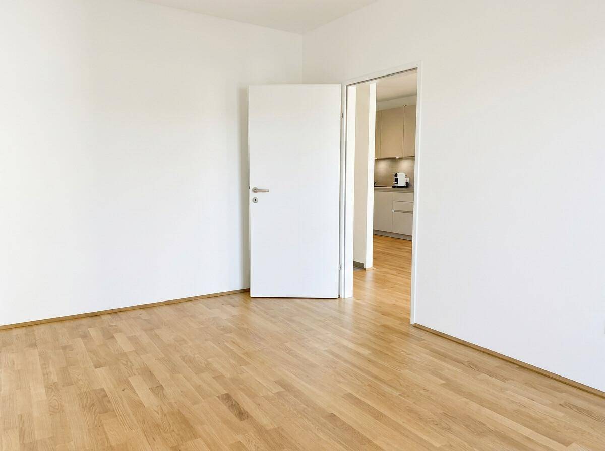 Leerer Raum mit hellem Parkettboden und einer offenen Tür, die einen Blick in eine moderne Küche gewährt.