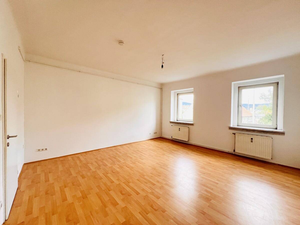 Geräumiges, leeres Wohnzimmer mit hellem Holzboden und zwei Fenstern.