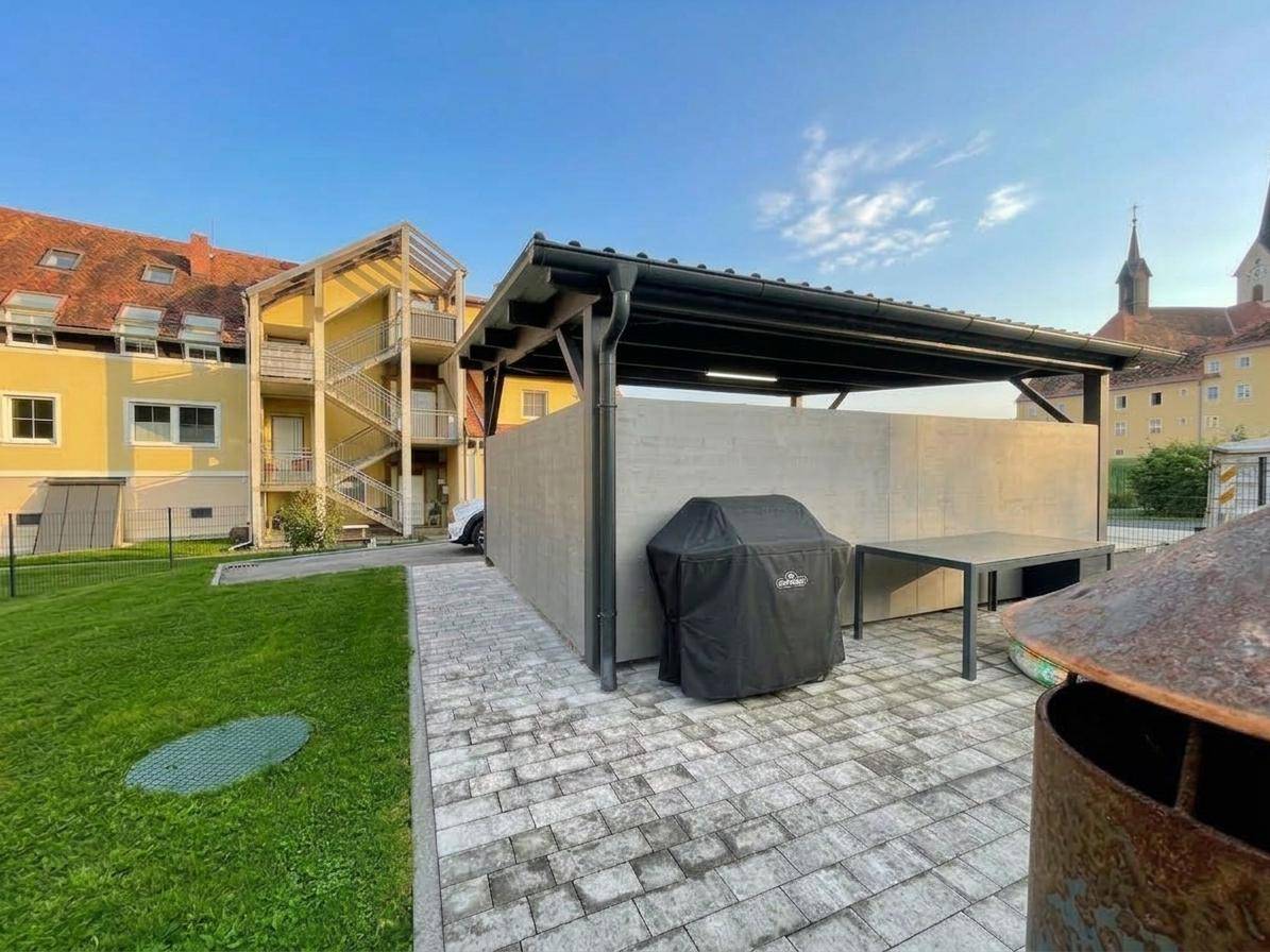 Außenbereich mit Grillplatz, überdachtem Sitzbereich und gepflegtem Rasen vor dem Gebäude.