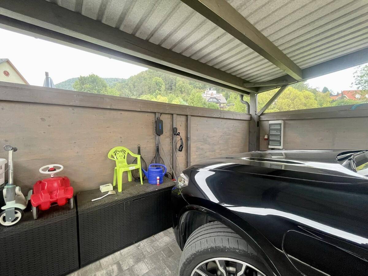 Überdachter Carport mit Elektroauto-Ladestation und Stauraum für Spielzeug, umgeben von Natur.