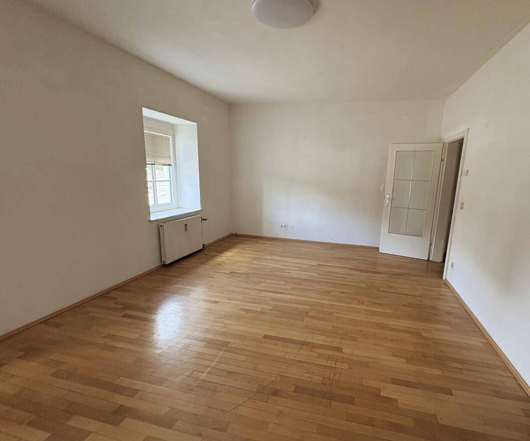 Leerer Raum mit hellem Parkettboden, weißen Wänden und einem Fenster, das natürliches Licht hereinlässt.
