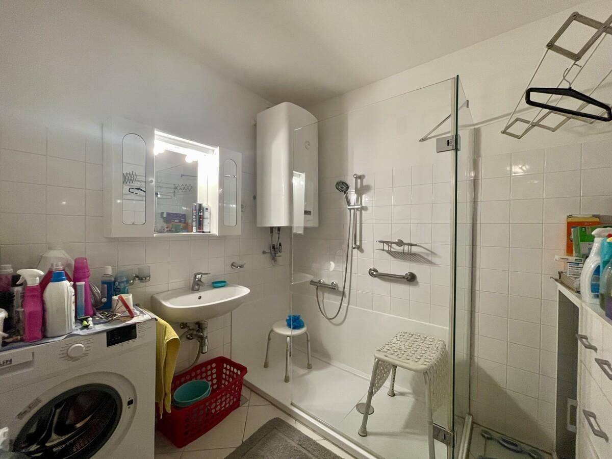 Helles Badezimmer mit weißen Fliesen, Dusche, Waschbecken und Waschmaschine, funktional eingerichtet.