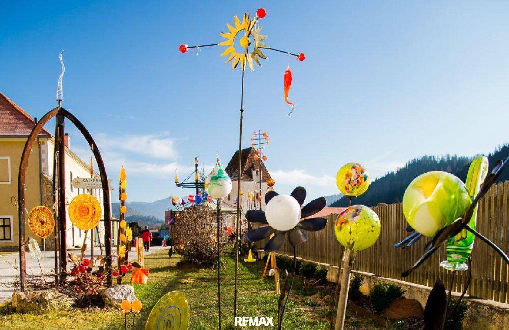 Künstlerische Gartendekorationen mit bunten Windspielen und Skulpturen in einer sonnigen Umgebung vor einem Gebäude.
