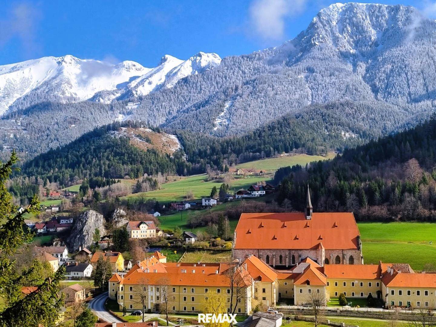 Historisches Klostergebäude in einem malerischen Dorf, umgeben von schneebedeckten Bergen und grünen Tälern.